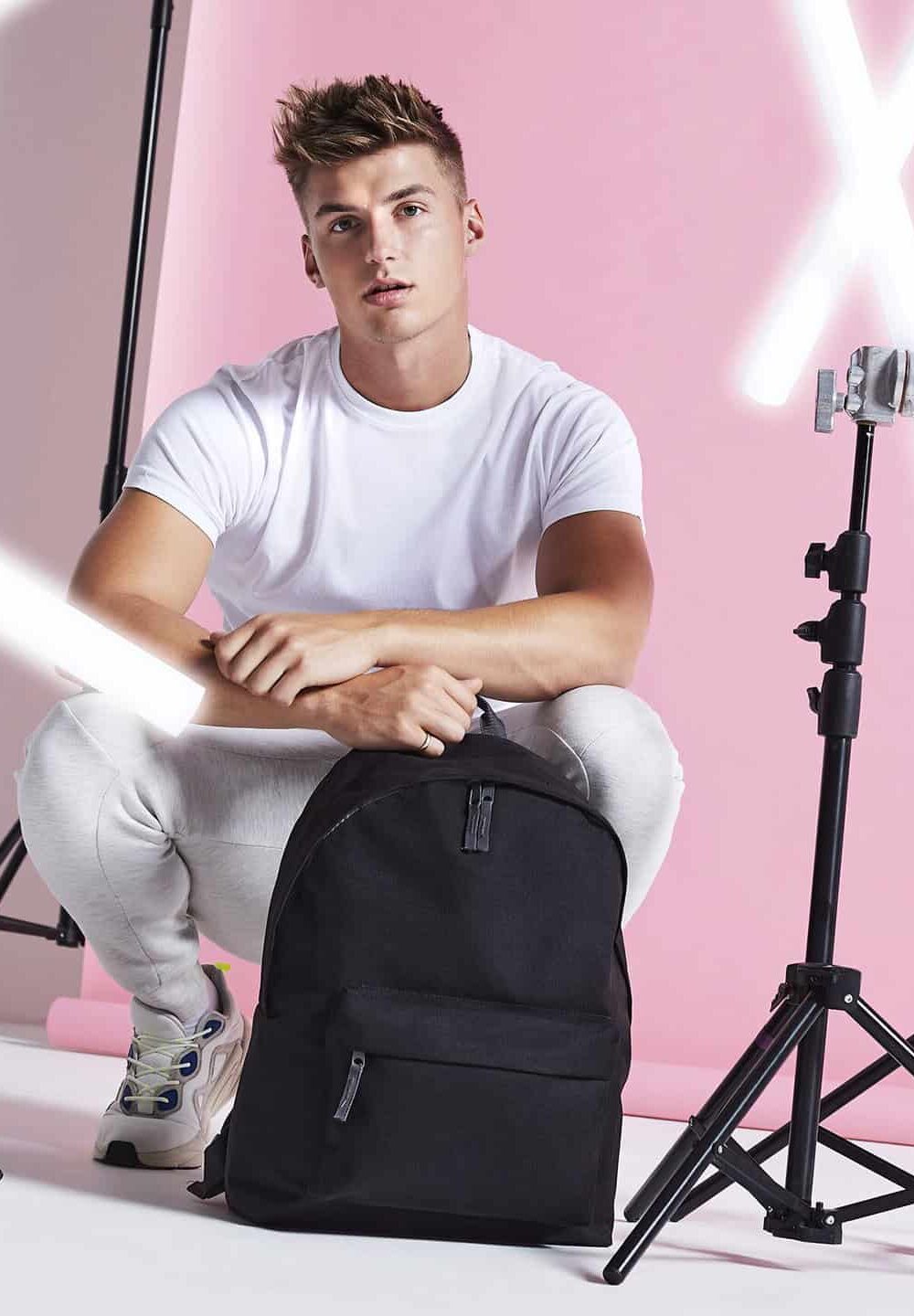 High school fashion model with casual white t-shirt, seated with black backpack, vibrant pink photography studio background, professional studio lighting, promoting style and youth fashion.