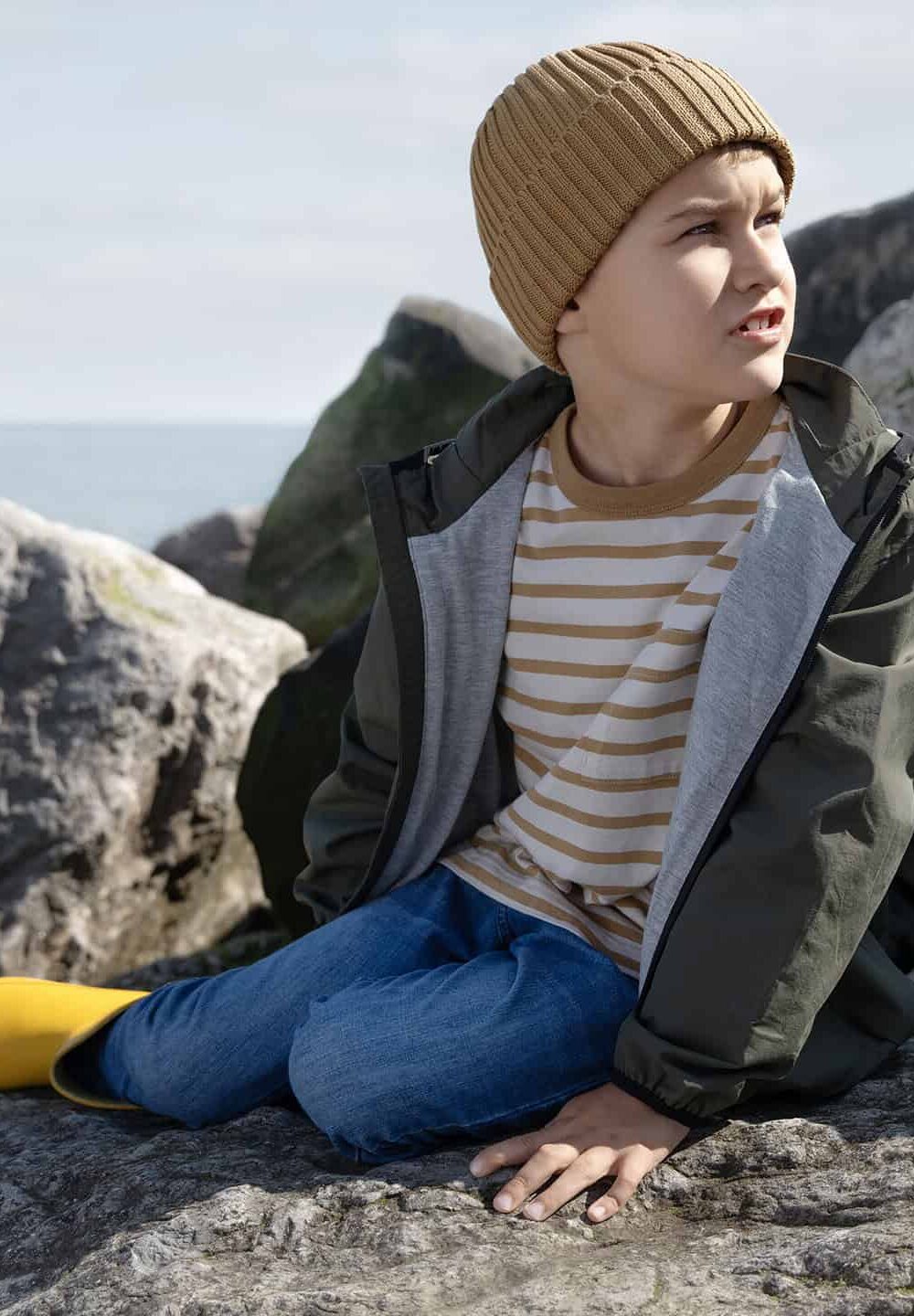 Boy sitting on rocks wearing outdoor jacket and yellow waterproof boots.