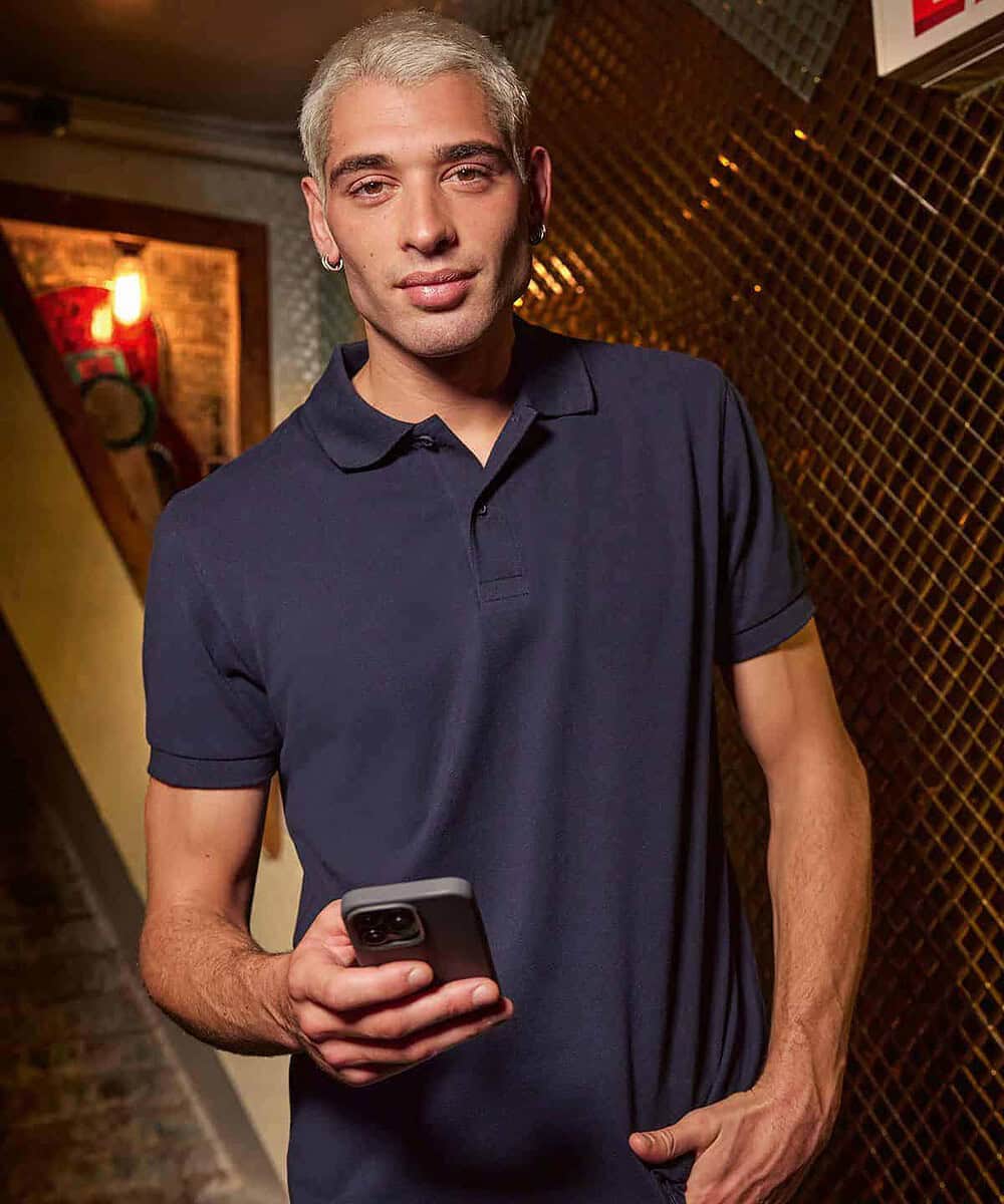 Man wearing navy B&C Eco Polo shirt, holding a smartphone in a stylish indoor setting.