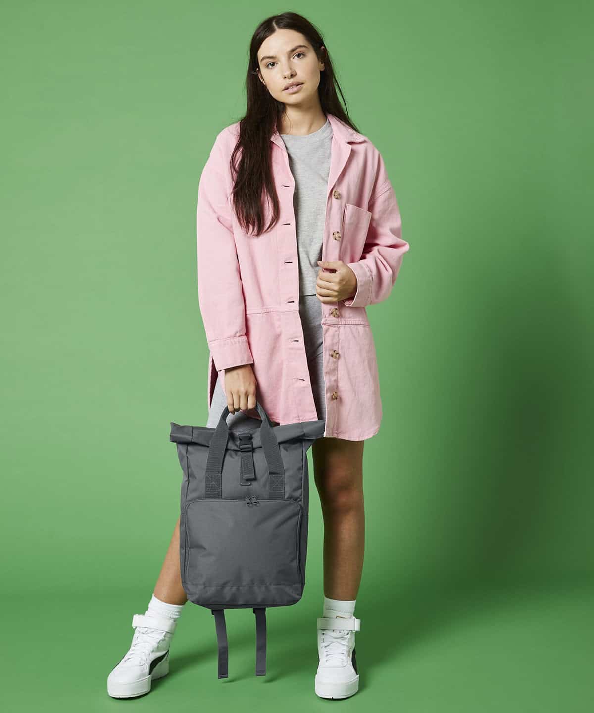 Stylish young woman in casual pink denim coat holding a grey backpack against a green background, showcasing personalised embroidery workwear and school uniform options.