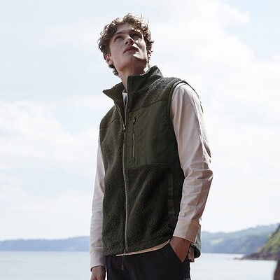Front view of a young man wearing a Recycled Sherpa Gilet on a beach background.