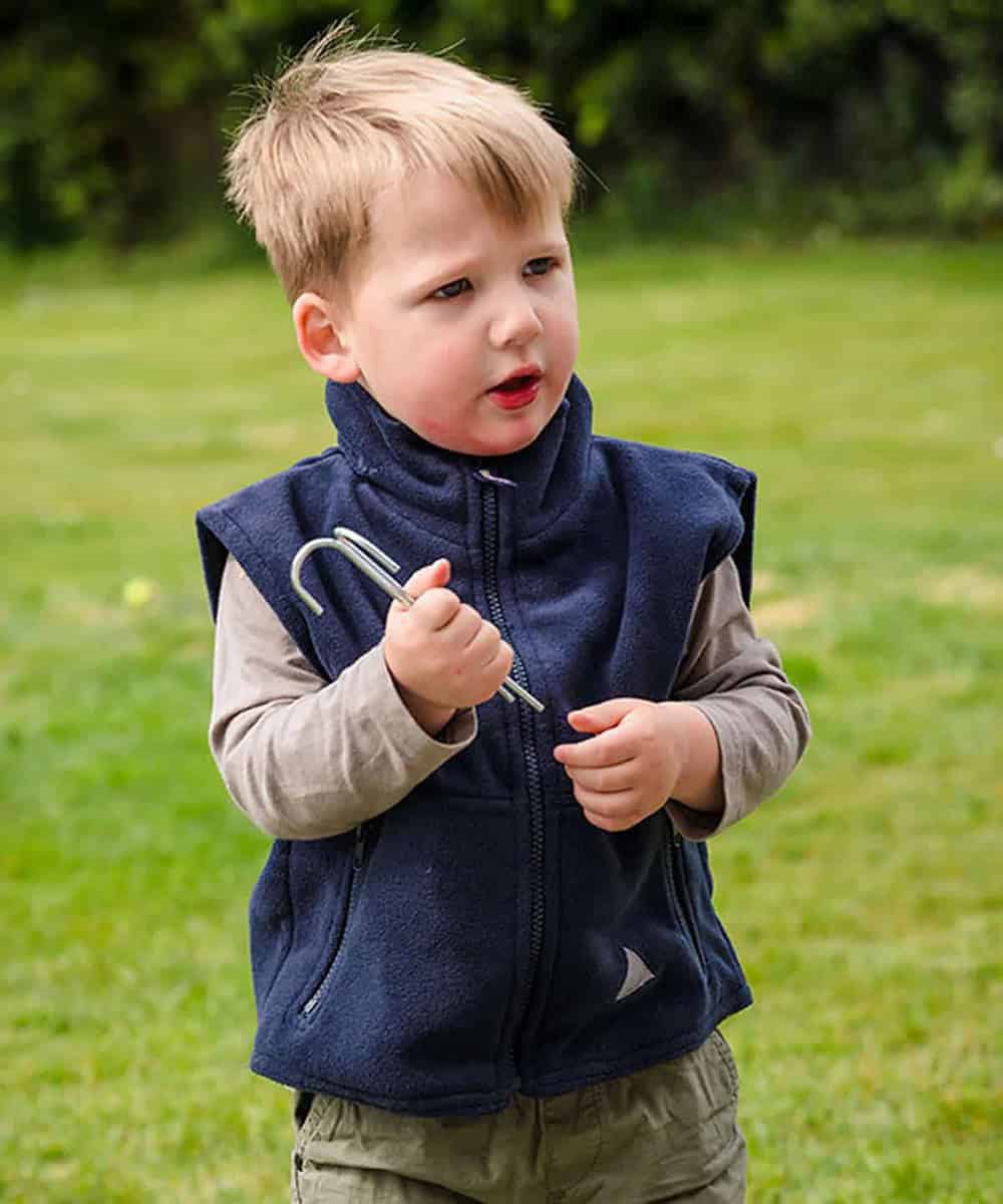 Child wearing a navy blue insulated vest outdoors, holding a small gardening fork.