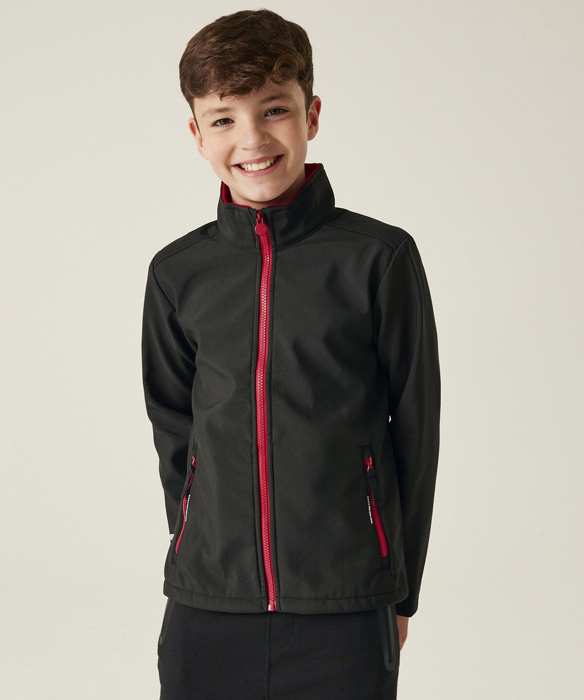 Youth black softshell jacket with red zippers, ideal for personalised workwear and embroidered uniforms, featuring a high collar and practical pockets. Perfect for casual or corporate branding.