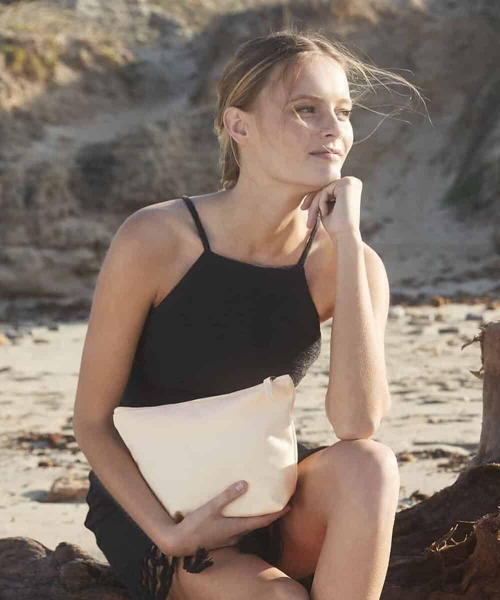 Woman holding a beige organic accessory bag on the beach, eco-conscious lifestyle.