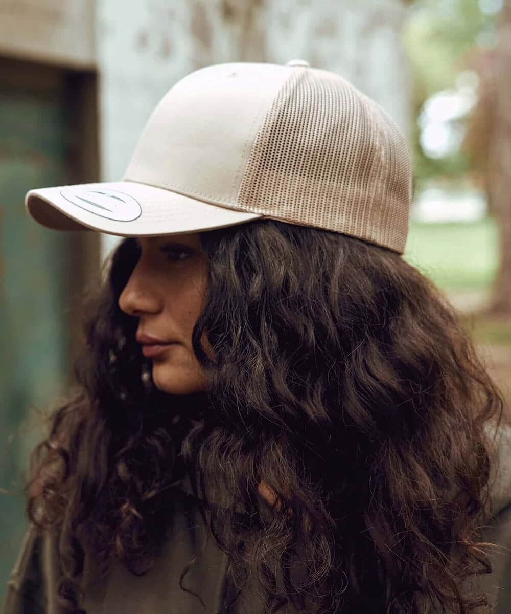 An image of a woman wearing a beige Yupoong YP023 retro trucker cap outdoors.